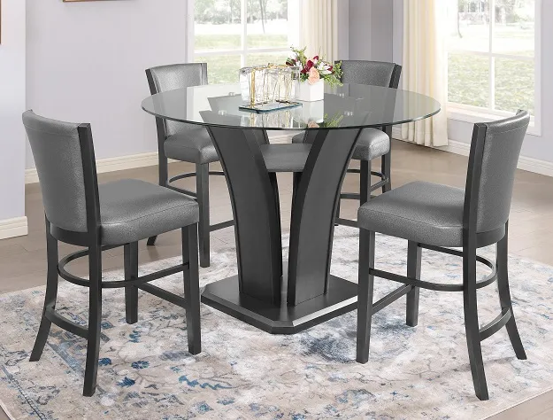 The "Camelia" Counter-Height Dining Set (#1716) in dark grey features a round glass table and four gray upholstered chairs, arranged on a rug in a bright room with large windows. A floral centerpiece and glasses decorate the table.