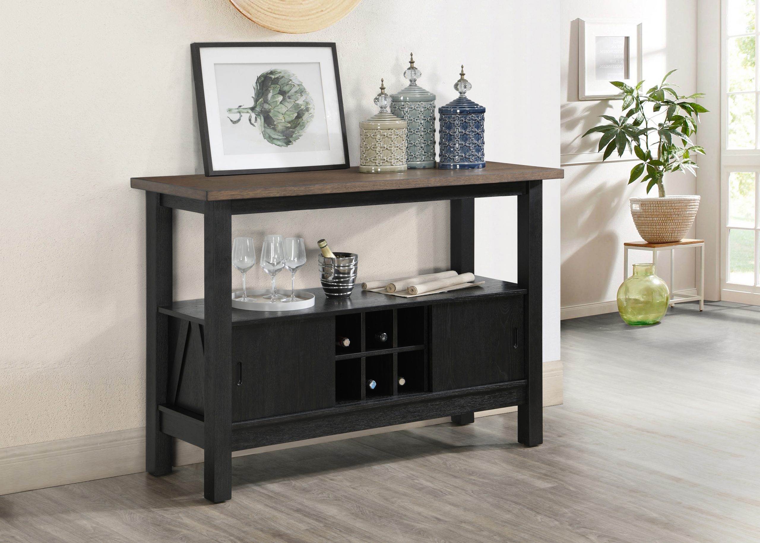 The Maribelle Server (#2158WC-SERVER) in Wheat/Charcoal displays glass jars, framed art, glasses, a wine bottle, and storage cubbies. A potted plant is visible near a sunlit window in the background.