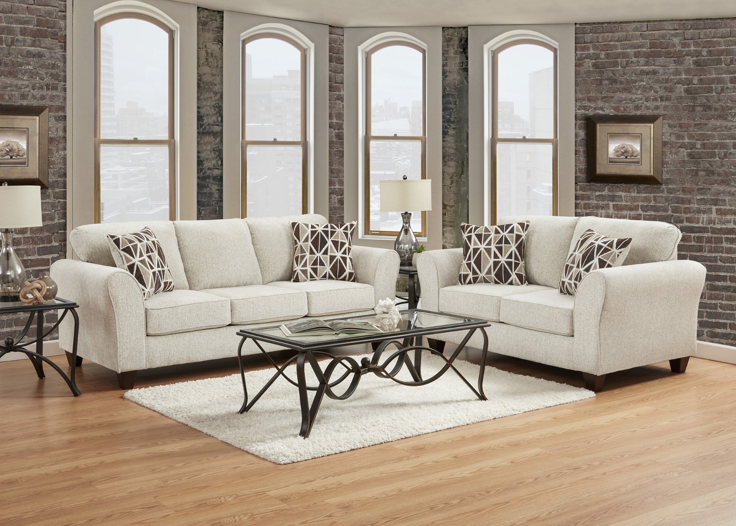 A modern living room featuring the #AF5000 - The Bita Buff Sofa & Loveseat Set in light gray, geometric pillows, a glass coffee table with metal frame, white rug, two table lamps, wooden floors, brick walls, and tall arched windows.
