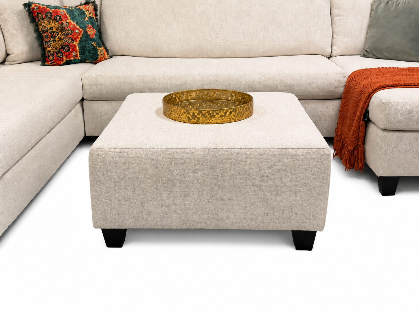 The #4250 Square Ottoman in beige sits before a matching sectional sofa, topped with a round gold tray, and is accented by a colorful pillow and an orange throw blanket.