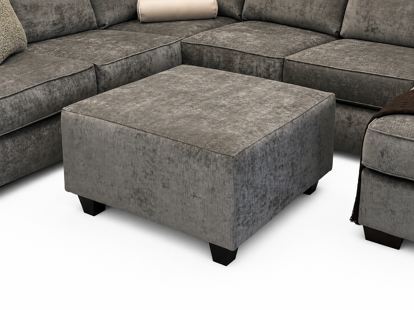 The #4250 Square Ottoman with dark legs sits on a white floor, surrounded by matching gray sectional sofas with cushions and a brown throw blanket.