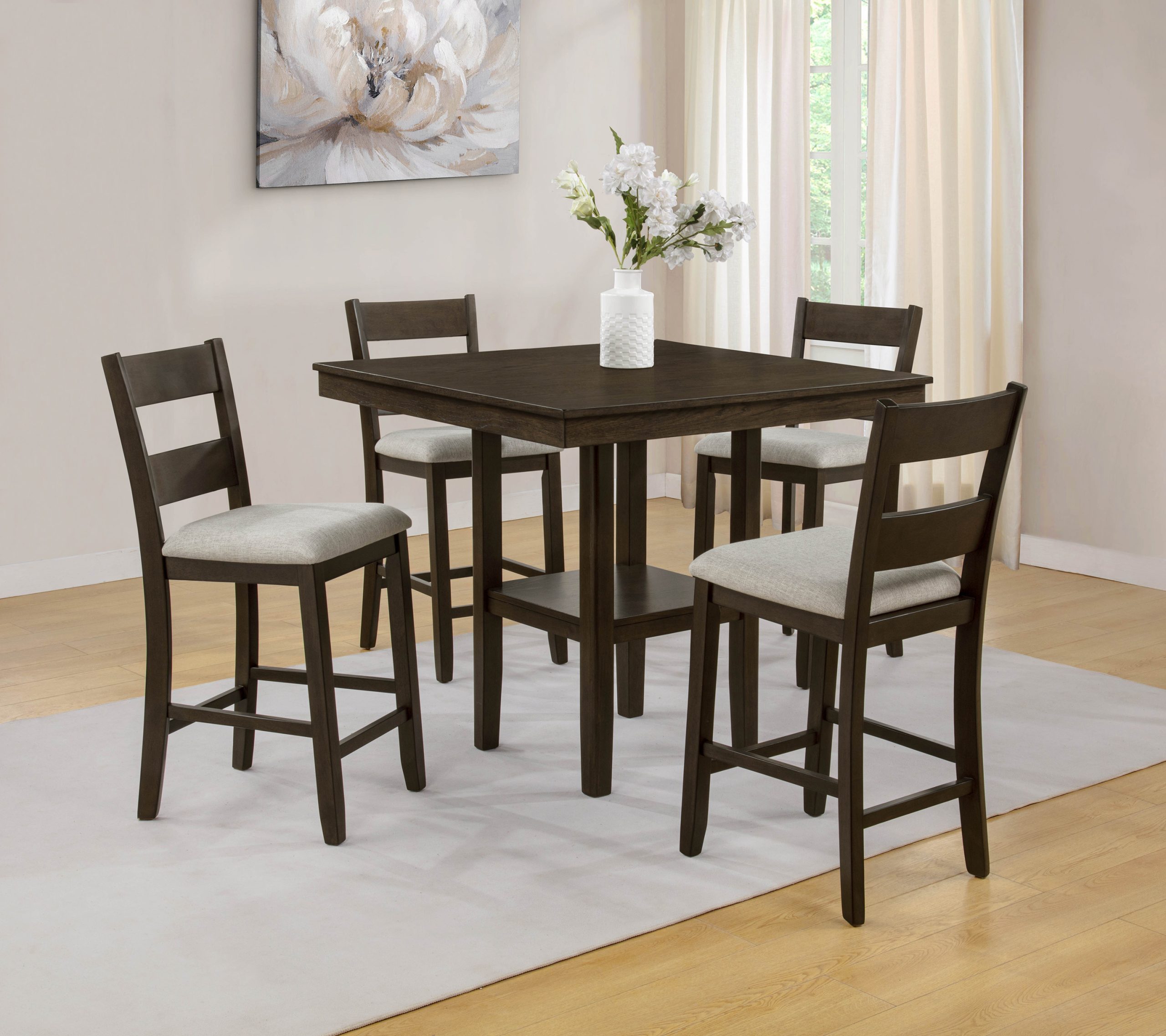 The "Lester" Counter-Height Dining Set in Brown (#2631BRN) features light cushioned seats on a brown table and chairs, set on a light rug in a bright room, accented by a white vase with flowers and a floral wall painting.