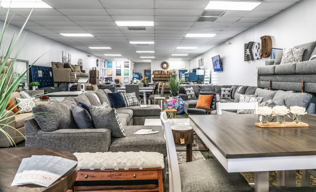 A furniture showroom featuring gray sofas, sectionals, and armchairs—perfect for discovering furniture design ideas for Charlotte homes. Dining tables with chairs and décor accents are showcased under bright ceiling lights along artfully decorated walls.
