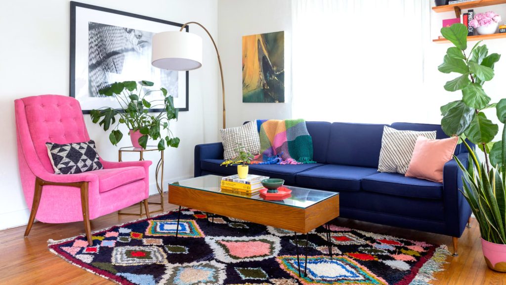 A colorful living room with a navy blue sofa, multicolored pillows and blanket, pink armchair, glass coffee table, patterned rug, lush houseplants, and framed art on sunlit white walls—perfect inspiration for any furniture buying guide Charlotte NC.
