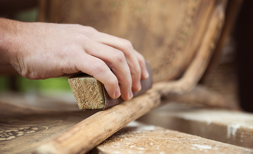 A close-up of a hand using a sanding block to smooth the edge of a wooden chair, with wood shavings visible below—a key step in furniture care and maintenance.
