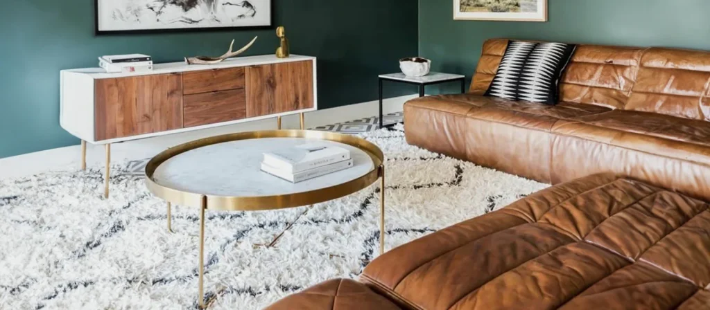 A modern living room with a brown leather sectional sofa, round marble coffee table, white shag rug, wooden sideboard with decor, and green walls adorned with framed artwork—designed for easy furniture care and maintenance.