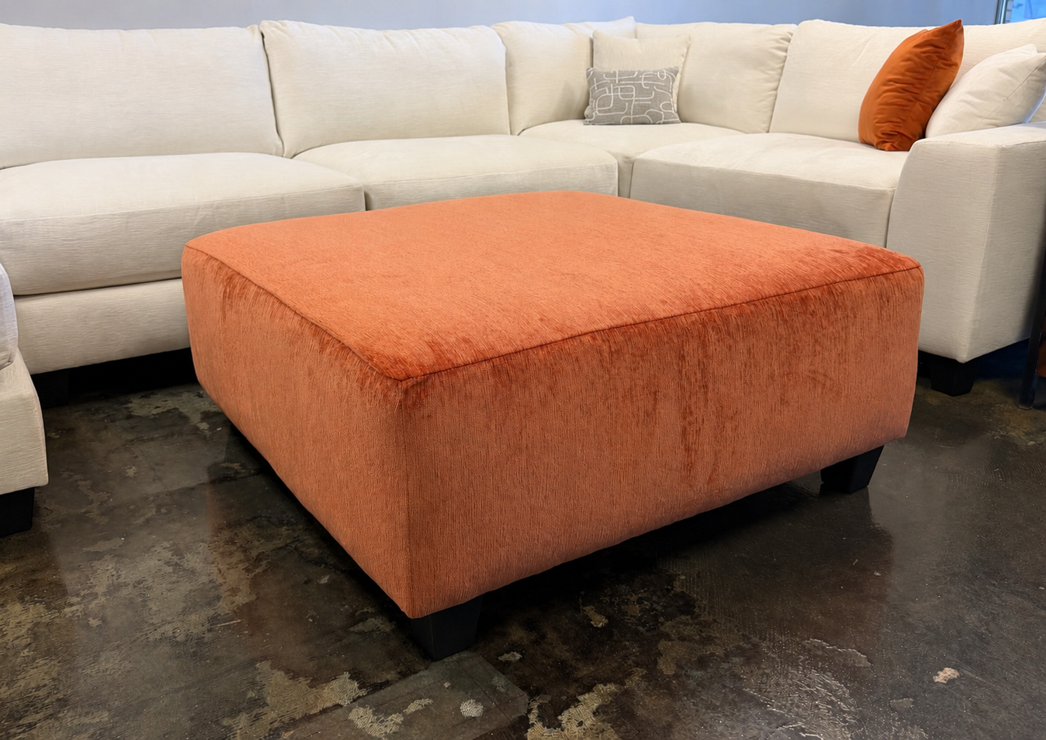 The #1099 Square Ottoman in orange rests on a polished concrete floor in front of a light beige sectional sofa decorated with stylish cushions.