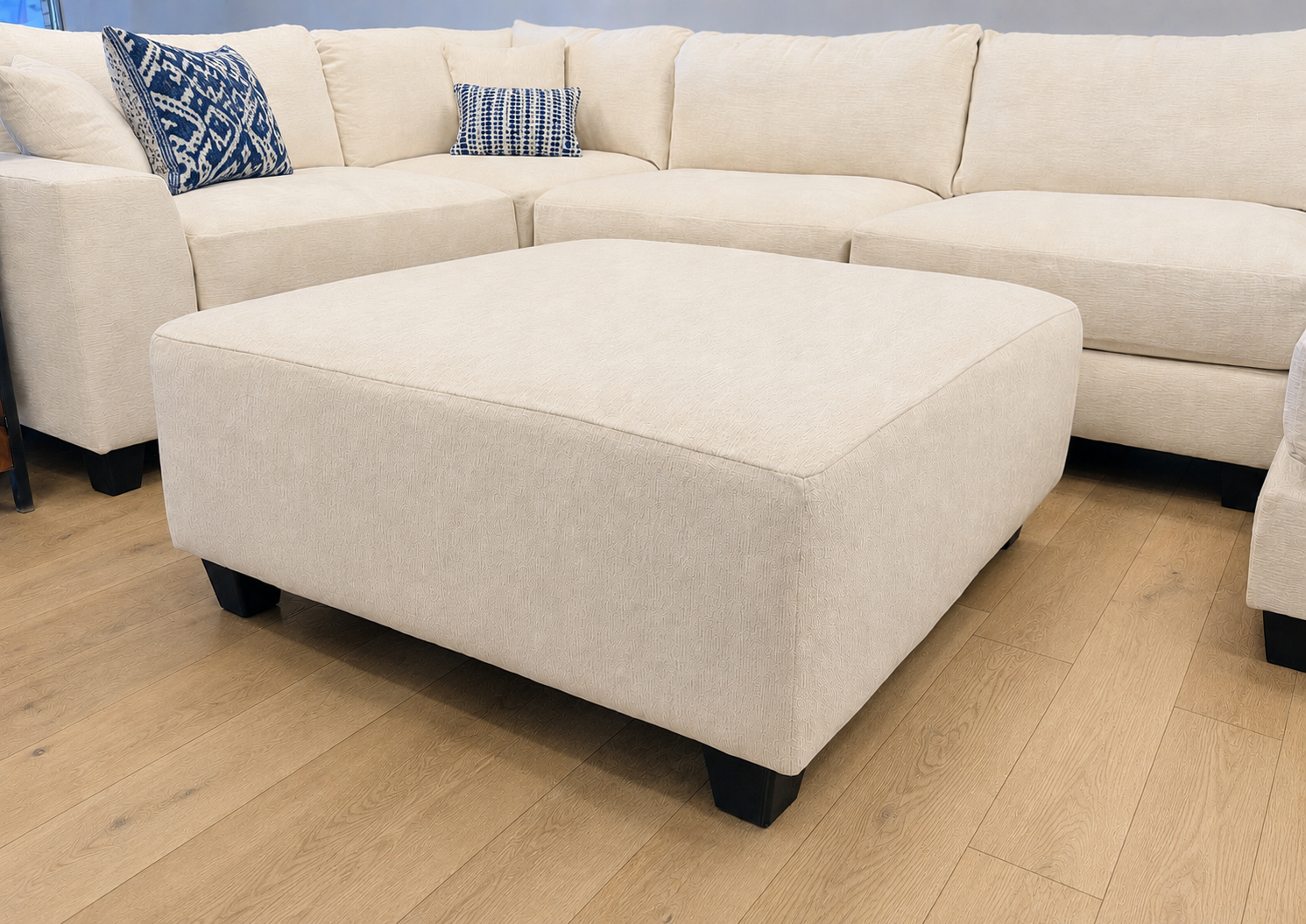 The #1099 Square Ottoman is placed on a light wooden floor in front of a matching L-shaped sectional sofa, accented with blue and white patterned pillows.